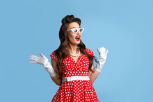 Stylish Pin Up Woman In Polka Dot Red Dress And Sunglasses Feeling Excited, Opening Mouth In Shock On Blue Background