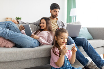 Gadgets addiction. Eastern family using different electronic devices, resting together in living room at home