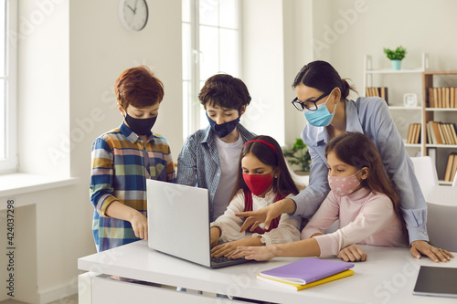 Primary school kids and teacher wearing medical face masks in computer lab. Happy junior students and tutor coding game on laptop. Basic programming skills, STEM education, flu epidemic, health care