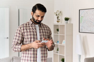 Wall Mural - Young 30s indian man holding smartphone tech device using cell phone apps at home. Bearded ethnic guy texting messages looking at smart phone checking social media, ordering online or browsing.