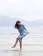 © Raisa Kanareva - Image of a young brunette woman in a blue dress walking barefoot on a beach and dangles his feet in the water