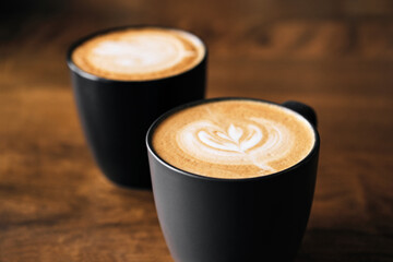 Wall Mural - Two cappuccino coffee with latte art on wooden table. Black cups and beautiful milk foam. Soft focus