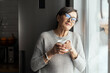 © Vadim Pastuh - Beautiful senior woman wearing eyeglasses enjoying morning coffee, stands in the kitchen at home. A calm middle aged woman with a mug of hot drink looks at camera