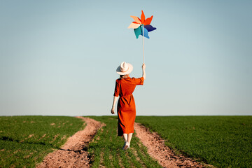 Wall Mural - Woman in a red dress with pinwheel on country road in green wheat field in spring