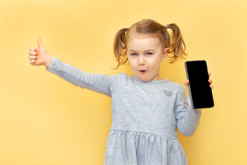 Excited cute child hold mobile phone with blank empty screen, showing thumb up and looking at camera with shocked face. Online shopping goods and service.Banner, place for text, mockup