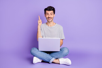 Photo of guy sit floor raise finger hold netbook shiny smile wear white t-shirt jeans sneakers isolated purple background