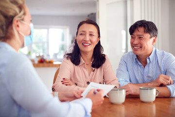 Smiling Mature Asian Couple At Home Meeting With Female Healthcare Worker In Mask During Pandemic