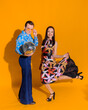 Front view of man and woman dressed in clothing like from 70s on the yellow wall isolated. Man holding a disco ball and couple posing for the photoshoot.