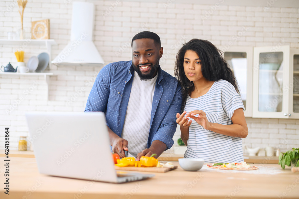 Interested young mixed-race couple attentively watching online cooking classes, learning how to make dinner lunch pizza, watching video blog course from laptop in the kitchen, preparing vegetables - obrazy, fototapety, plakaty 