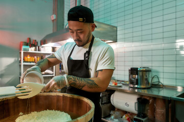 Wall Mural - Young male chef cook adding vinegar while making sushi rice in a wooden barrel in the kitchen of Japanese restaurant