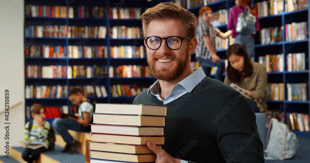 Bearded school teacher or librarian holding books in hands standing in library interior. - obrazy, fototapety, plakaty 