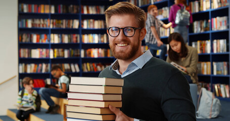 Canvas Print - Bearded school teacher or librarian holding books in hands standing in library interior.