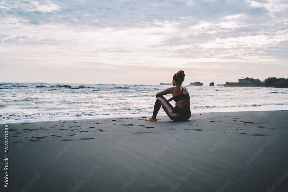 Slim woman resting at black sand of seashore thinking about cardio training for have muscular body, back view of female runner in sportive clothing resting during morning workout at coastline - obrazy, fototapety, plakaty 