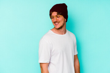 Young caucasian man wearing a wool hat isolated on blue background  looks aside smiling, cheerful and pleasant.