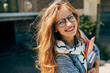 © iuricazac - Candid portrait of a happy young student woman with long red hair smiling and wearing transparent eyeglasses standing next to the unversity and carrying lots of books and folders on a sunny day.