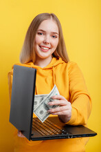 Beautiful Young Woman Freelancer Takes Out Dollars Money Banknotes From Laptop. Win In Online Casino Game Wealth By Freelancer Work. IT Remote Work Via Laptop Isolated Over Yellow Color Background.