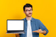 © Kateryna - Handsome charismatic stylish caucasian guy, holding an open laptop with a blank white screen in his hand, points with his hand at it, stands on isolated orange background, place for your presentation