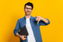 Unhappy Caucasian Stylish Handsome Guy With Glasses, Freelancer Or Student, Holds A Laptop In Hand, Shows A Thumb Down Gesture, Stands On Isolated Orange Background, Displeased Facial Expression