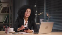 Portrait Of African American Woman Waiting For An Answer And Looking At Smartphone And Laptop Monitor. Businesswoman Posing At Workplace, Sitting At Table In Office. Slow Motion Ready 59.94fps.