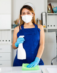 Young mexican woman in medical mask cleaning table with spray at company office