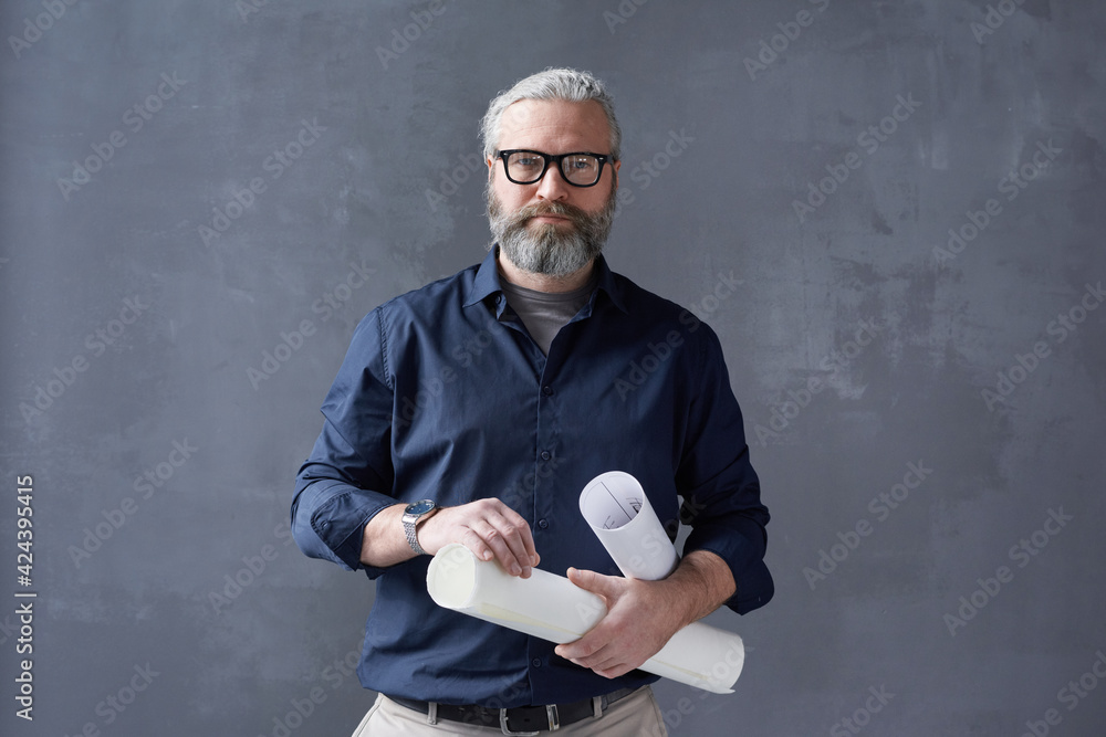 Portrait of mature bearded architect in eyeglasses standing with blueprints against the black background - obrazy, fototapety, plakaty 