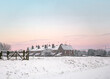 Beautiful winter sunset over row of old houses in countryside with deep snow and glowing sky. Snowy scene as sunrise at dawn pink orange skies. Gate and wooden fence in foreground.