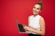 © Ivan Traimak - Side profile photo shot of beautiful smiling blond young woman with gathered hair wearing white t-shirt holding computer laptop typing on keyboard looking at camera isolated over red wall background