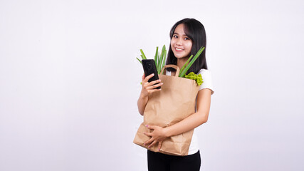 asian woman with paper bag of fresh vegetables and holding a phone with isolated white background