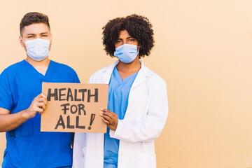 African American woman and Caucasian Hispanic young man dressed as medical staff holding a banner that reads 