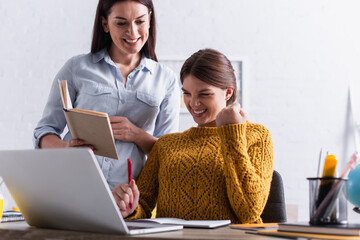 Wall Mural - excited teenage girl holding pen while doing homework near laptop and mother with book