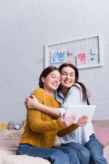 Wall Mural - cheerful mother hugging smiling teenage girl with digital tablet