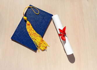 Canvas Print - Yellow tassel from graduation cap, blue diploma and paper scroll tied with red ribbon with bow on beige wooden background, Flat lay, top view