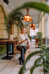 Wall Mural - Young business woman sitting in cafe at table, leaning his hand back in chair, looking out window and holding smartphone.