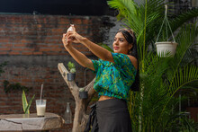 Portrait Of Beautiful Young Latin Woman With Long Black Hair, Sunglasses Over Head And Casually Dressed Taking A Selfie With Her Smart Phone