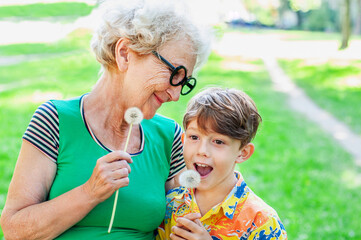 Wall Mural - Happy grandmother and grandchild hold dandelions pretending to be singing into microphone. Older woman, boy fooling around while on the background of a green lawn. Family relationships. Fun in spring