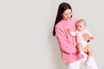 Dark haired Caucasian mother holding baby in hands and looking at her daughter, beautiful female dresses rose sweatshirt with infant isolated over white background with copy space.