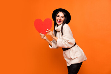 Sticker - Photo of young beautiful charming positive smiling girl hold big red paper heart present isolated on orange color background