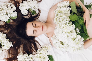 Close up of ?ute charming nice pretty woman lying on cozy bed with fresh spring flowers. Head shot portrait dreamy young brunette girl with eyes closed. Hair Care, skin beauty health care concept