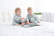 © Юля Шевцова - Two toddler baby twin boys in pajamas reading book sitting on white bedding on bed.