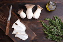 Three Raw King Oyster Mushrooms, Rosemary Sprigs, Mushroom Slices On A Cutting Board, Olive Oil On A Dark Wooden Background.