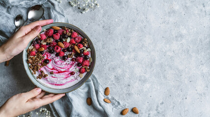 Hands holding healthy breakfast bowl with yogurt, granola, muesli, seeds, frozen raspberries, blueberries, nuts on light background. Healthy food, detox smoothy, vegetarian nutrition, top view