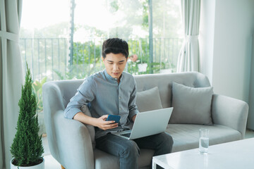 Wall Mural - Smiling young man typing on mobile phone while sitting on a sofa at home with laptop computer