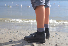 The Boy's Legs In Jeans Shorts, Socks And Dirty Shoes On The Seashore With Seagulls.