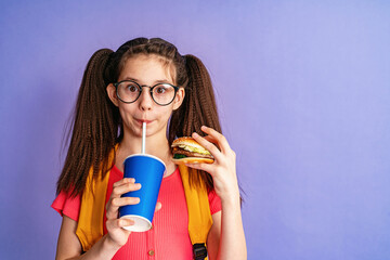 hungry schoolgirl with glasses and two ponytails drinks drink from a straw of a paper cup, holding hamburger purple background. child loves fast food. Fast food concept for school children. Copy space