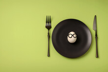White Egg With Glasses On A Black Plate, A Table Knife And A Fork On A Green Background. Easter Holiday Creative Concept. Flat Lay. Copy Space