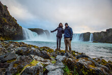 A Couple At The Bottom Of The Godafoss Waterfall With Sunset In The Background, Iceland