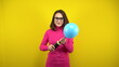 © Vital9c - A young woman inflates a blue balloon with a pump on a yellow background. Girl in a pink turtleneck and glasses.