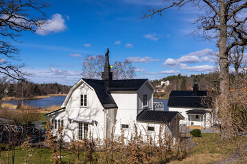 Old wooden two storey white house in traditional style. Black roof. Cottage on the lake shore. Spring season landscape. Sweden countryside in April. View of beautiful  European garden design. 