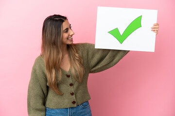 Wall Mural - Young caucasian woman isolated on pink background holding a placard with text Green check mark icon with happy expression
