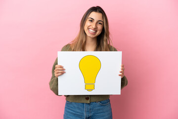 Wall Mural - Young caucasian woman isolated on pink background holding a placard with bulb icon with happy expression